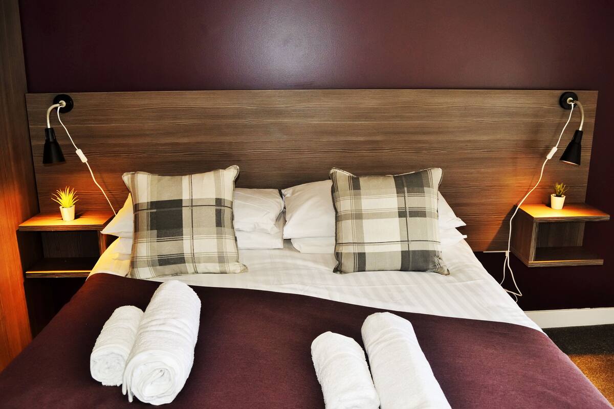 A neatly made bed with plaid pillows, a wooden headboard, and bedside tables with lamps and a potted plant.