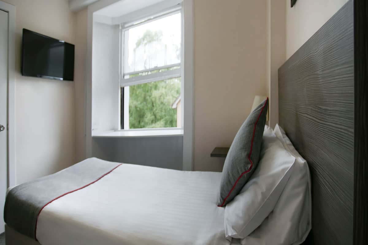 A neatly made bed with white bedding and red trim, a window with a view of trees, and a flat-screen TV mounted on the wall.