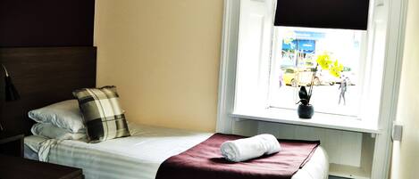 A single bed with a plaid pillow, a dark wooden headboard, a window with a view of the street, and a dark-colored blanket.