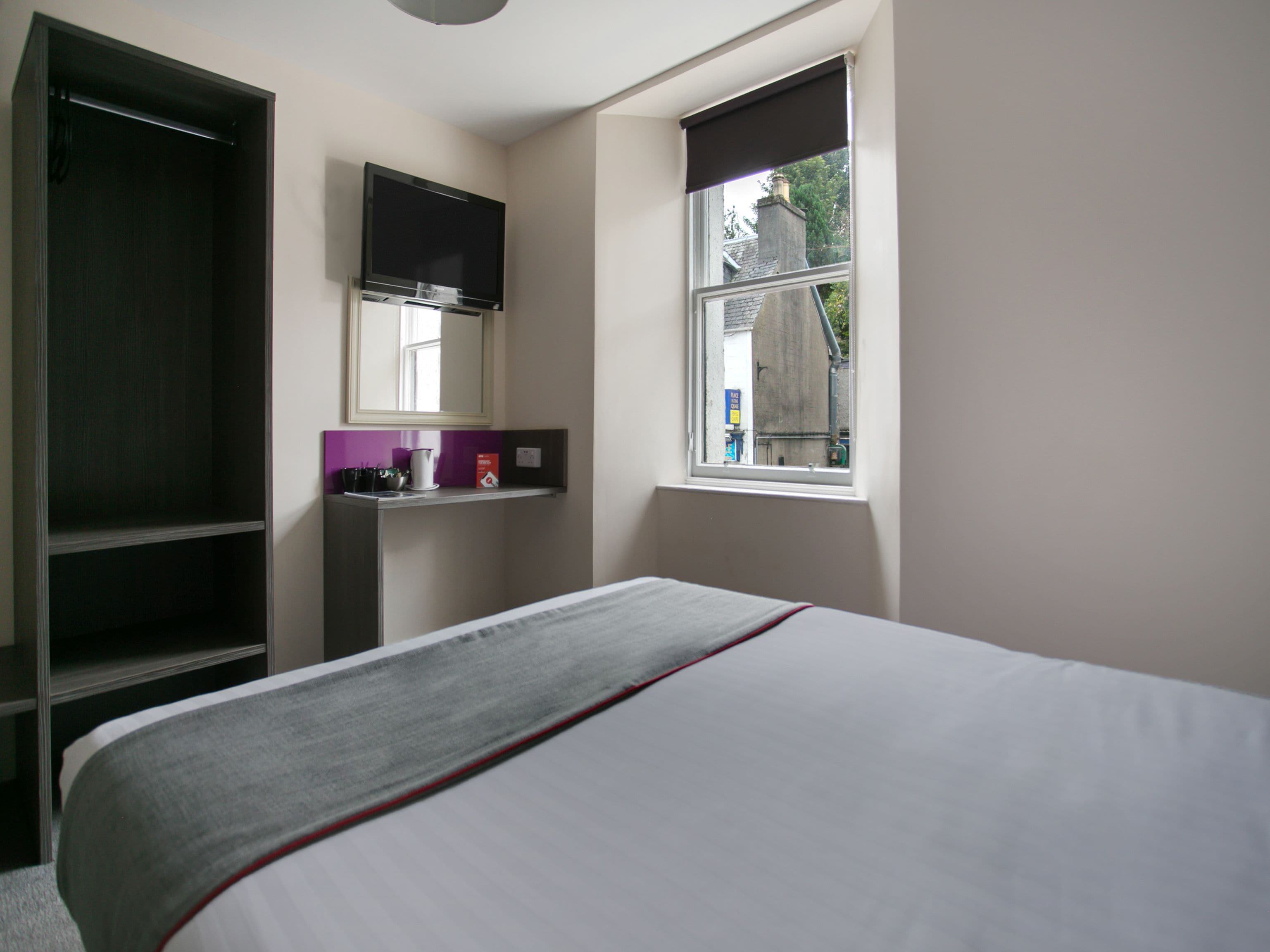 Un dormitorio con una cama, un televisor montado en la pared, una ventana y una estantería con objetos.