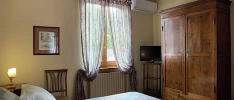 Un dormitorio con una cama, un armario de madera, un televisor sobre un mueble, una silla y una ventana con cortinas.