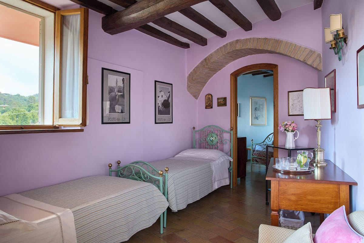 A room with two single beds, a wooden desk with a lamp, and a view of a blue door leading to another room.