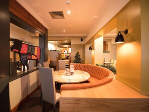 A modern restaurant with a round table and orange cushioned seating, a Christmas tree, and a menu board with food items. - Premier Inn Glasgow - Motherwell (Motherwell)