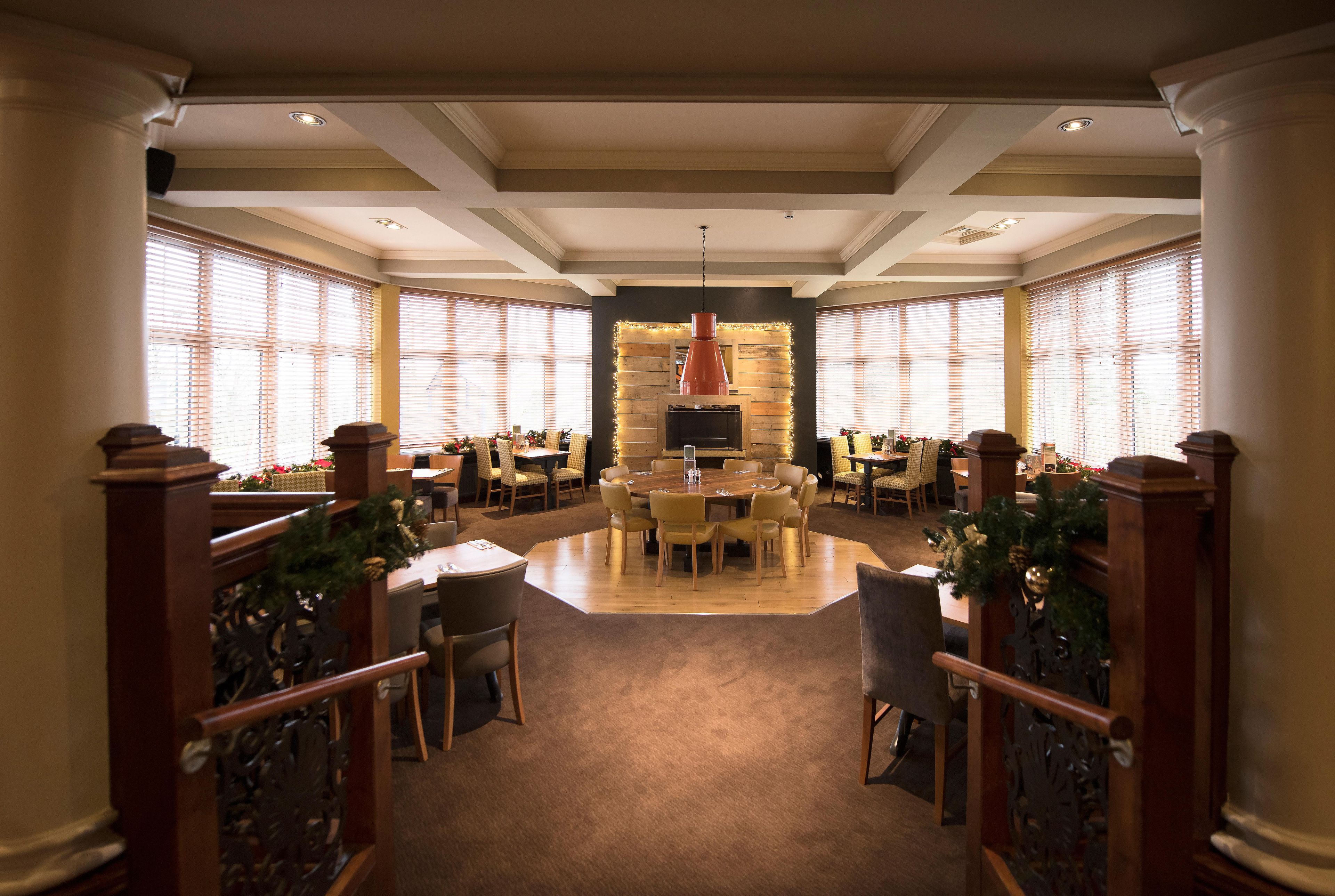 A spacious dining area with a central round table surrounded by chairs, flanked by large windows with blinds.
