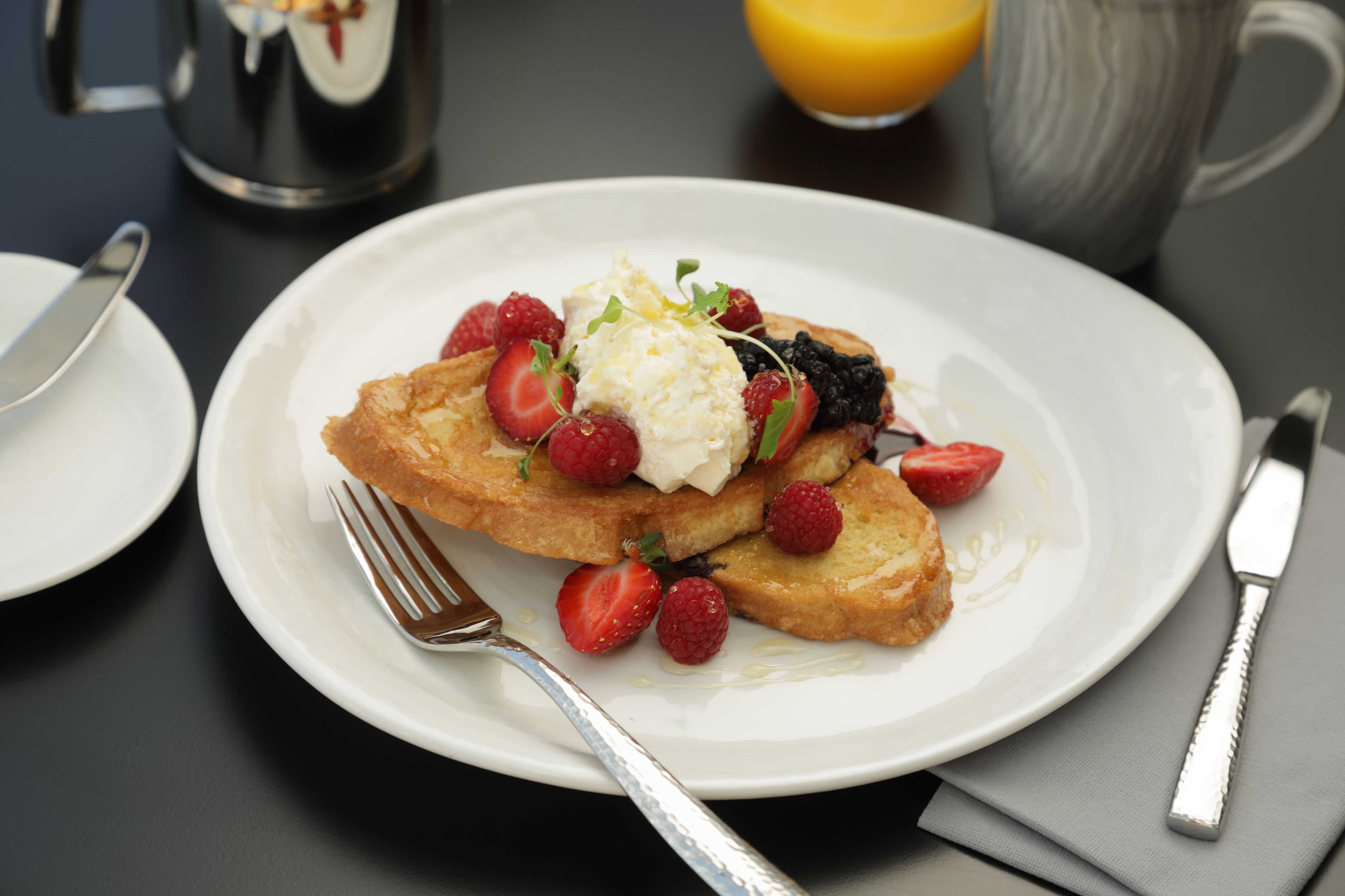A plate of French toast with strawberries and raspberries, a glass of orange juice, and a coffee pot on a table.