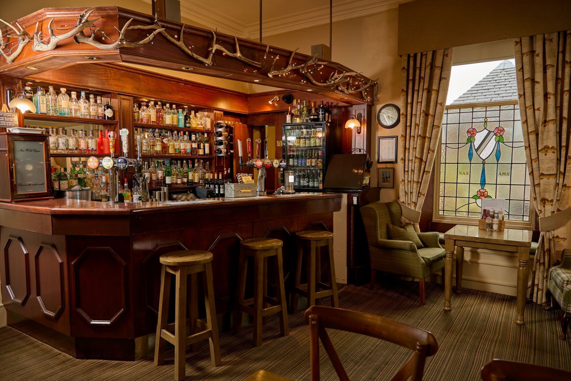 Un espace bar doté de tabourets en bois, d’une variété de bouteilles d’alcool et d’un élément décoratif en forme de bois de cerf placé au-dessus du bar.