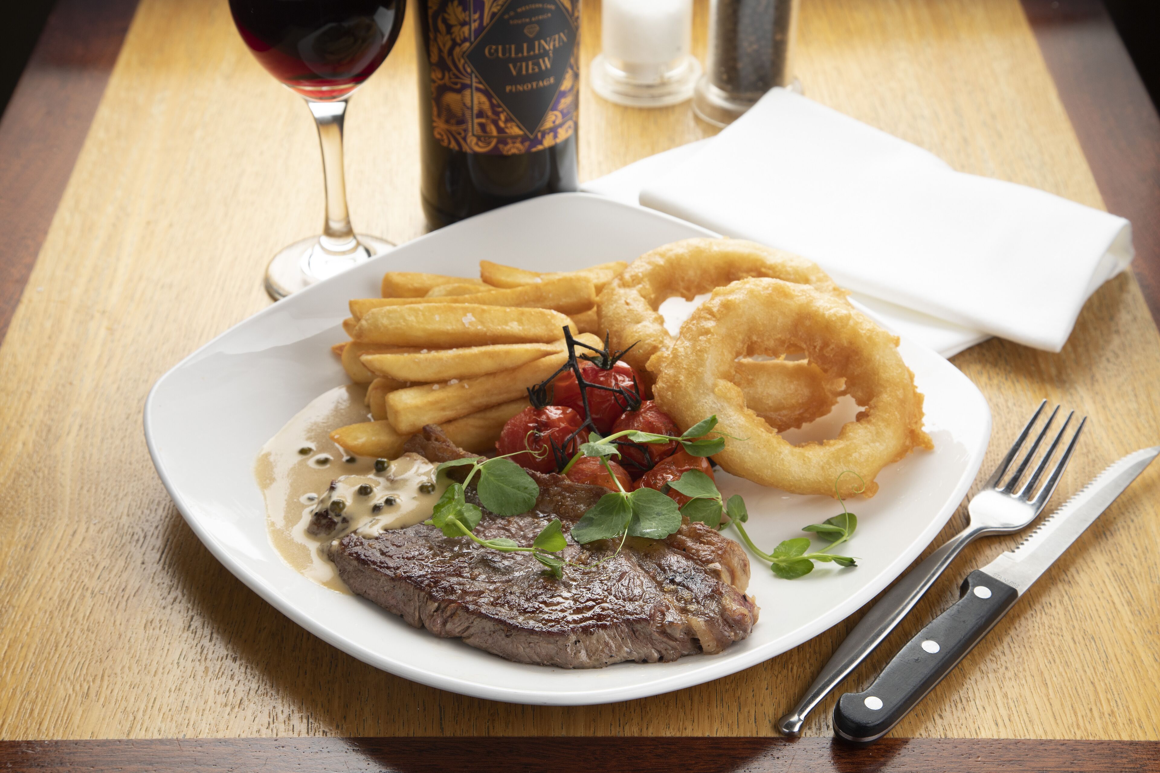 Un plato de comida con bistec, papas fritas, aros de cebolla y una bebida.