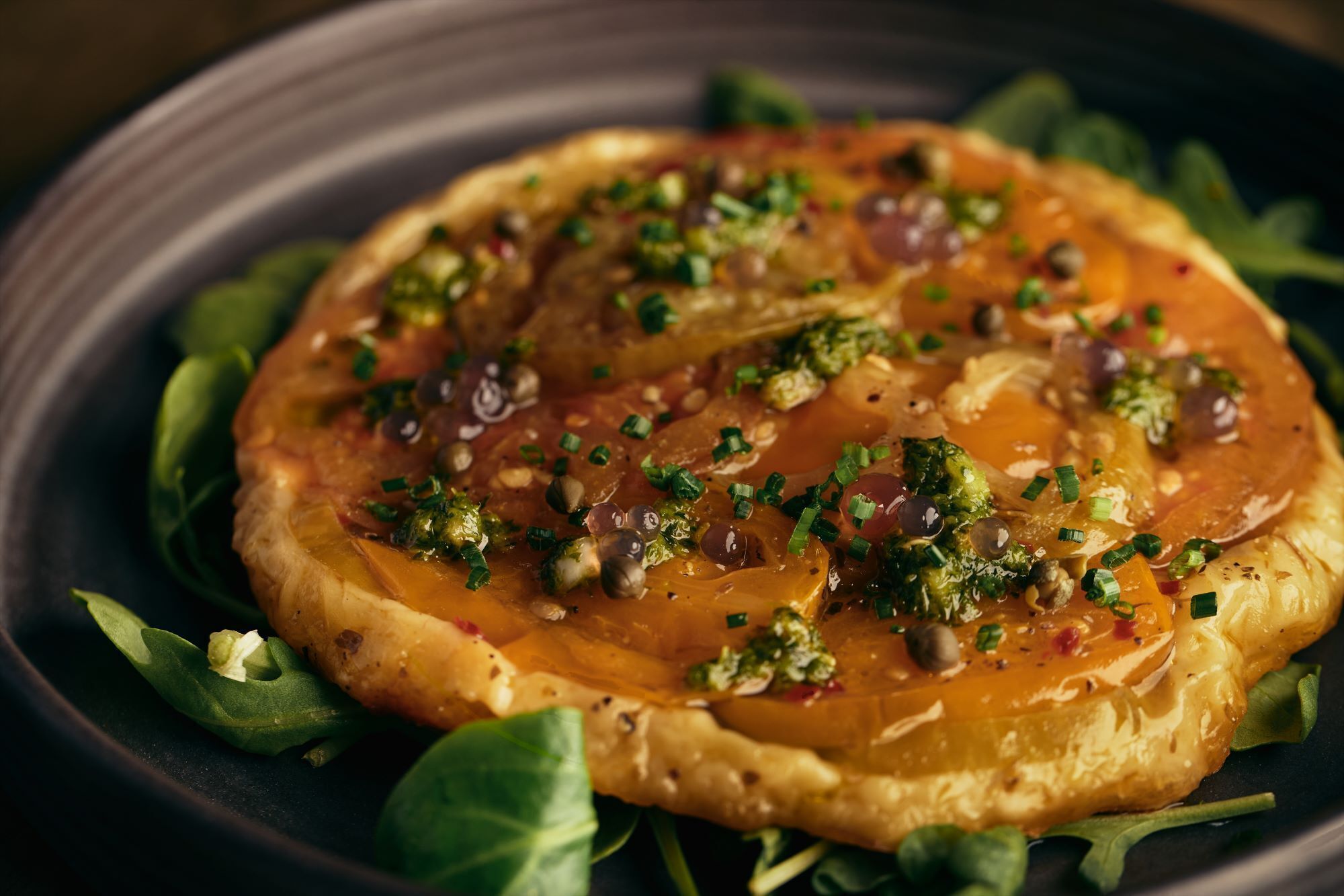 Un plat rond, doté d’une croûte dorée, garni d’herbes vertes et de petits éléments ronds, servi sur une assiette sombre, avec des feuilles vertes en dessous.