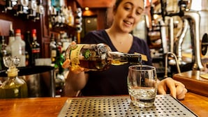 En bartender hælder en drink op i et glas i baren, med diverse flasker og et fad i baggrunden.