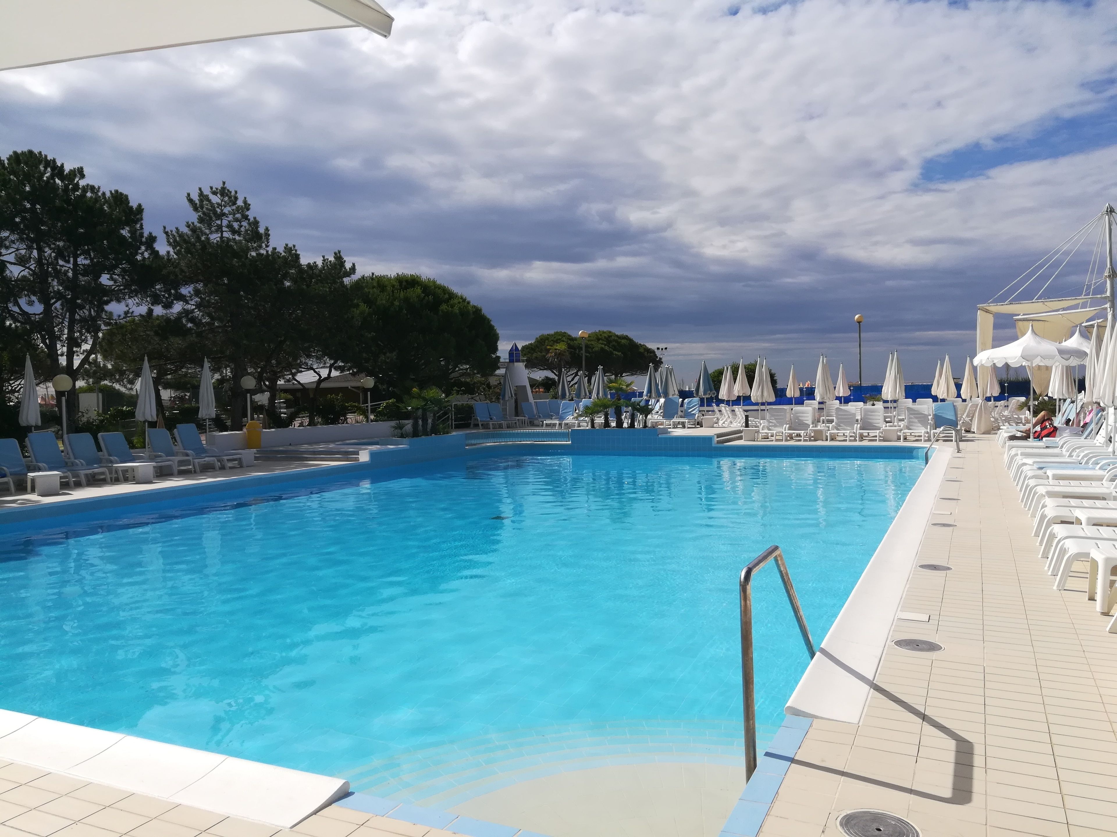 Una piscina con lettini e ombrelloni, circondata da alberi e un cielo nuvoloso.