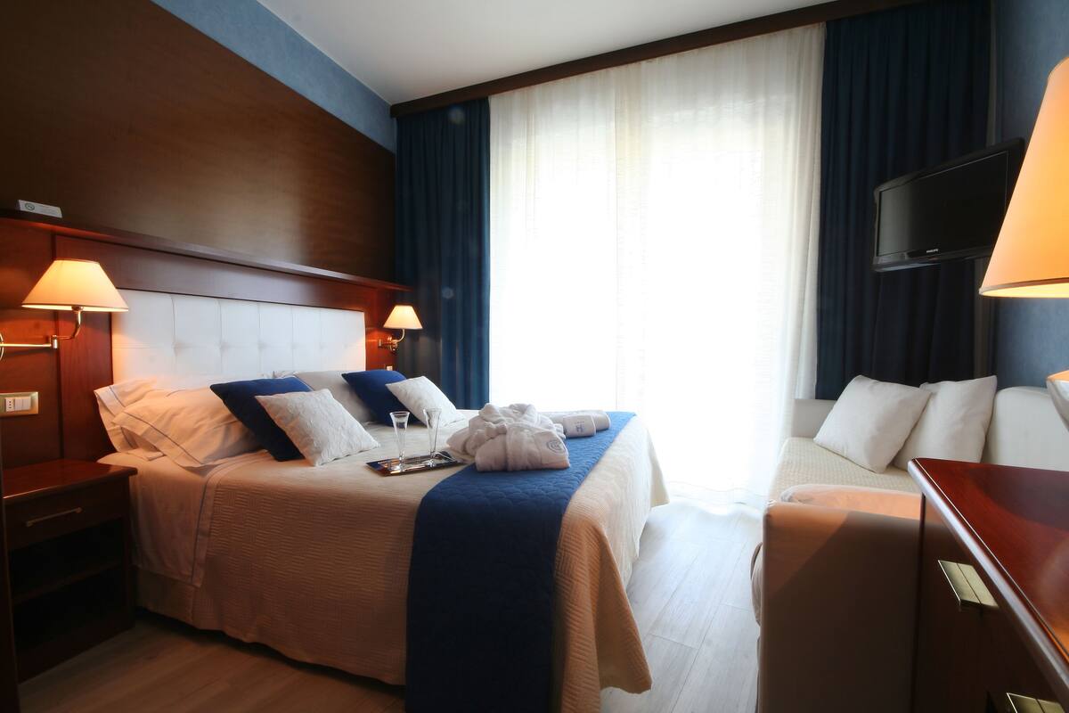 Une chambre d’hôtel avec un grand lit, des tables de chevet, une télévision et une fenêtre avec des rideaux.