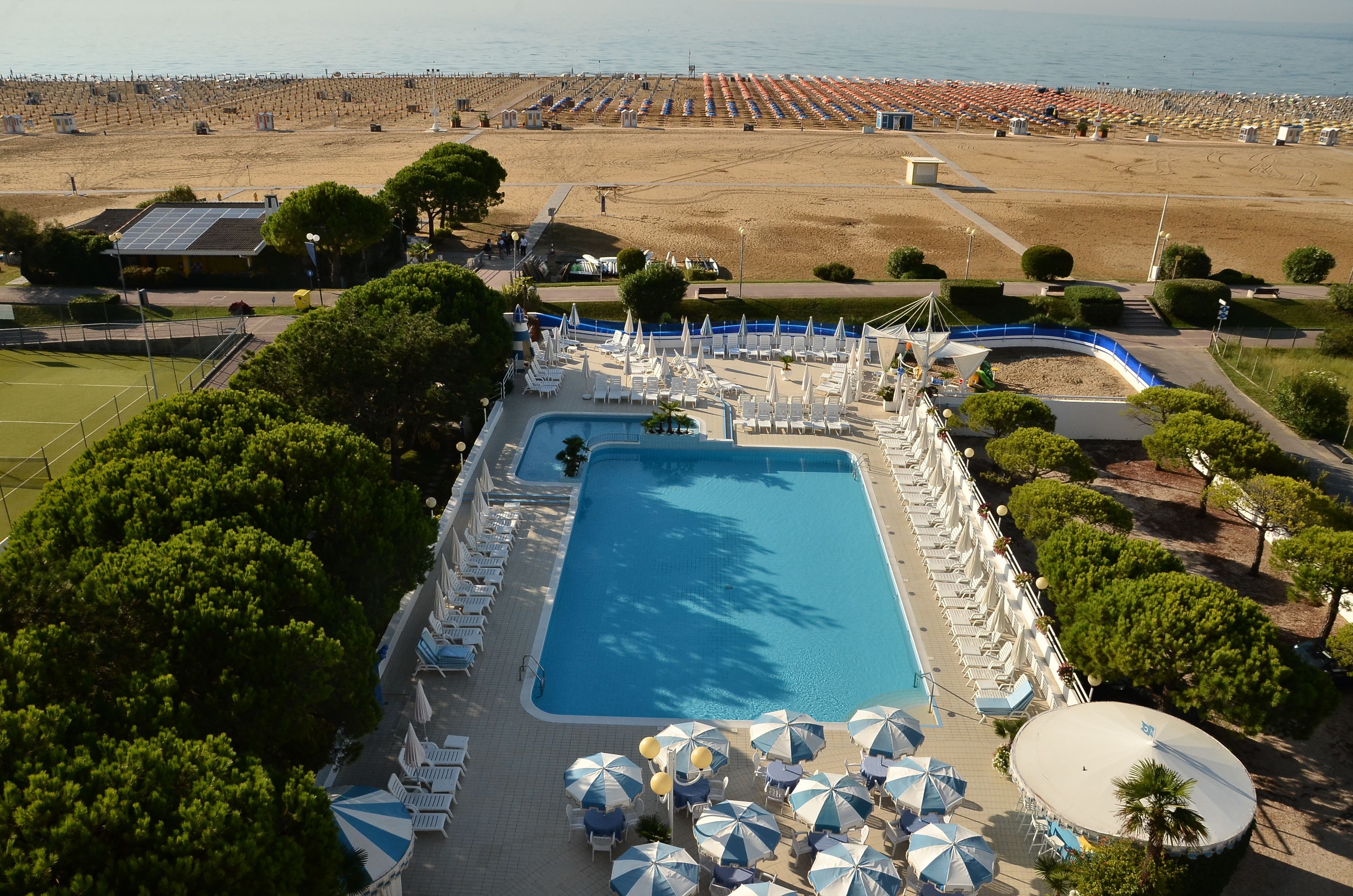 Ein Schwimmbecken mit Liegestühlen und Sonnenschirmen, umgeben von Bäumen und einem Strand im Hintergrund.
