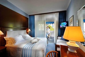 A hotel room with a bed, a desk, a chair, a TV, and a balcony with a person sitting.