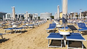 Ein Strand mit Liegestühlen und Sonnenschirmen, feiner Sand und Gebäude im Hintergrund.
