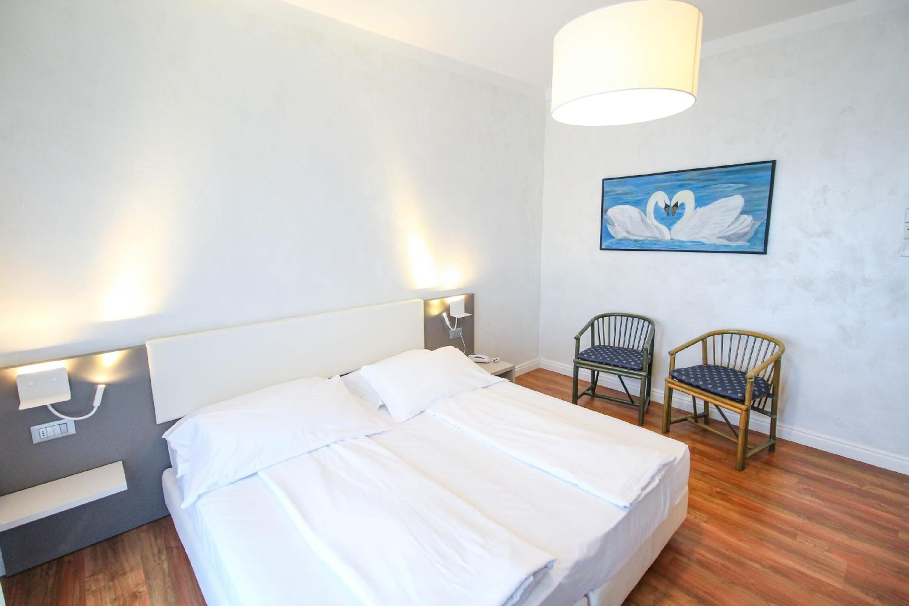 Habitación de hotel con cama, dos sillones, un cuadro de cisnes y una lámpara en la pared.