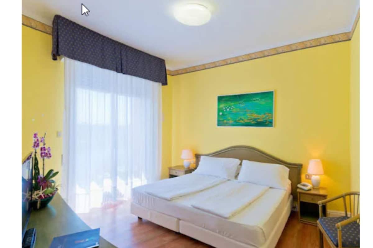 A hotel room with a bed, two bedside tables with lamps, a chair, a desk with a computer, a window with curtains, and a painting on the wall.