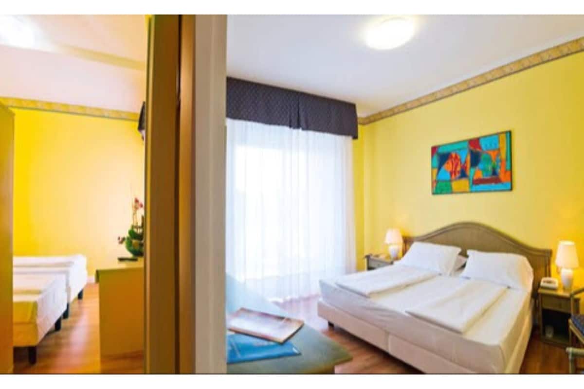 A hotel room with two beds, a large window with sheer curtains, a painting on the wall, and a bedside table with a lamp.