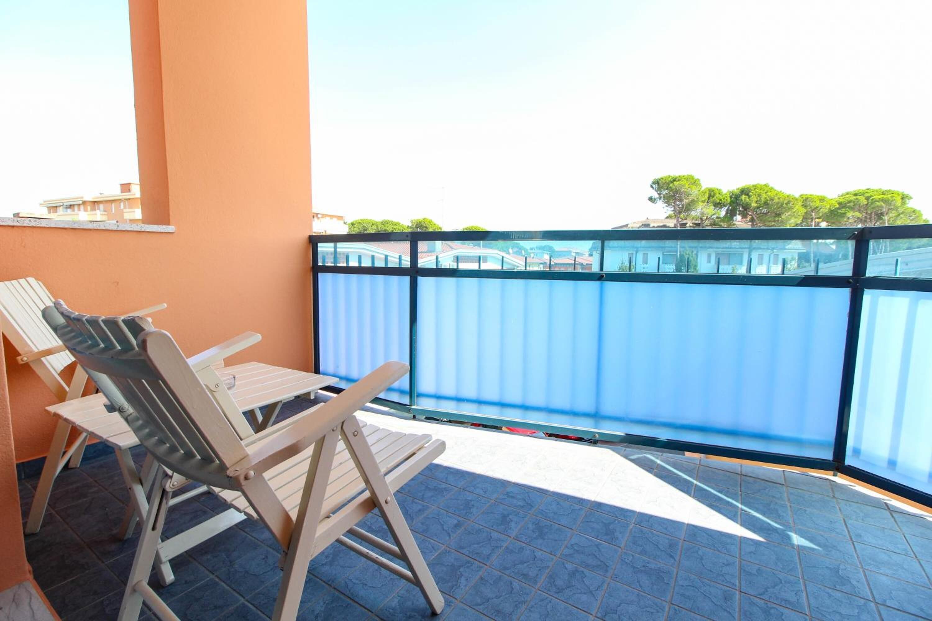 A balcony with two wooden chairs and a table, a blue railing, and a view of buildings and trees in the background.