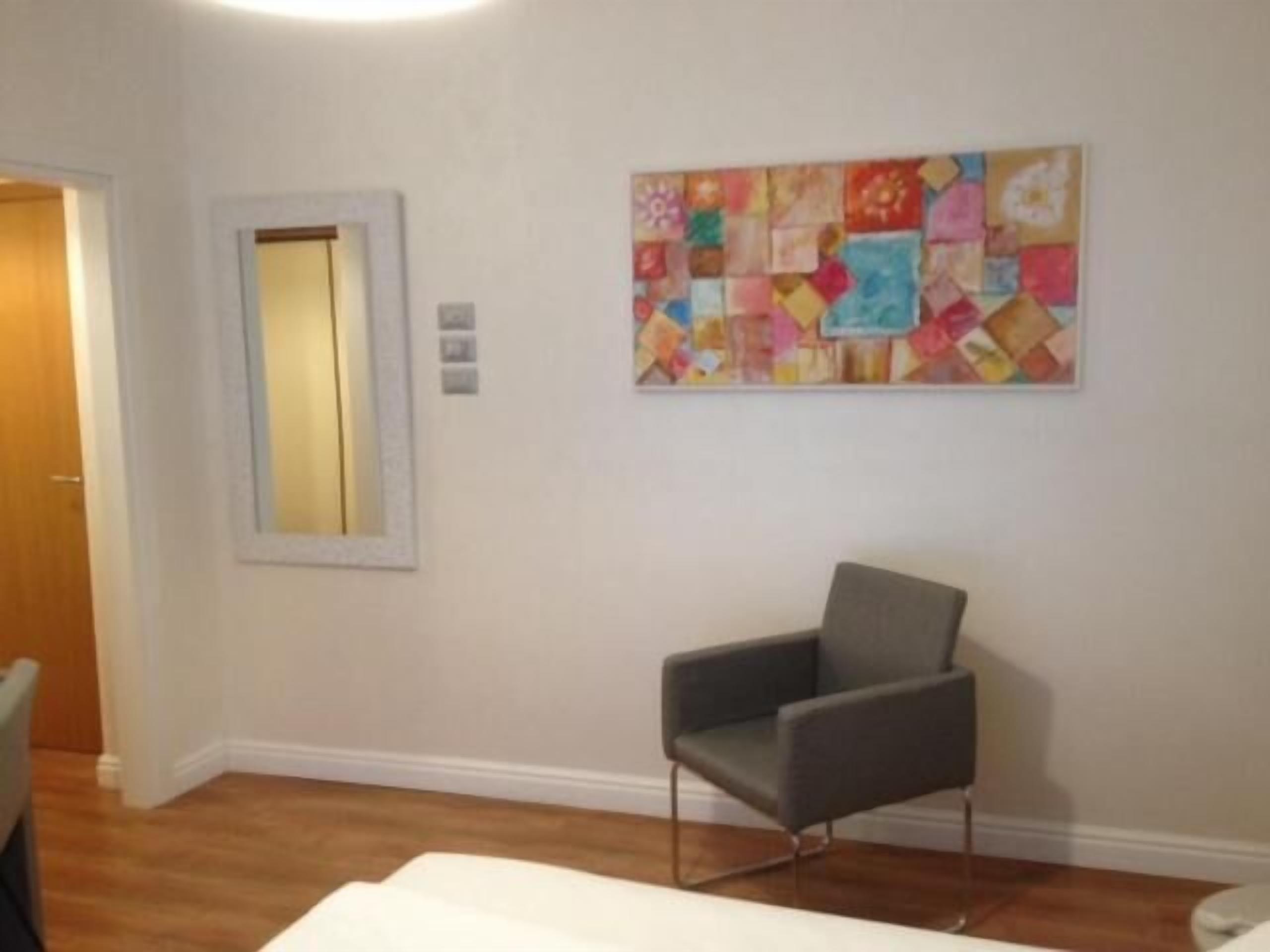 A room with a grey armchair, a colorful abstract painting on the wall, a wooden floor, and a white door.