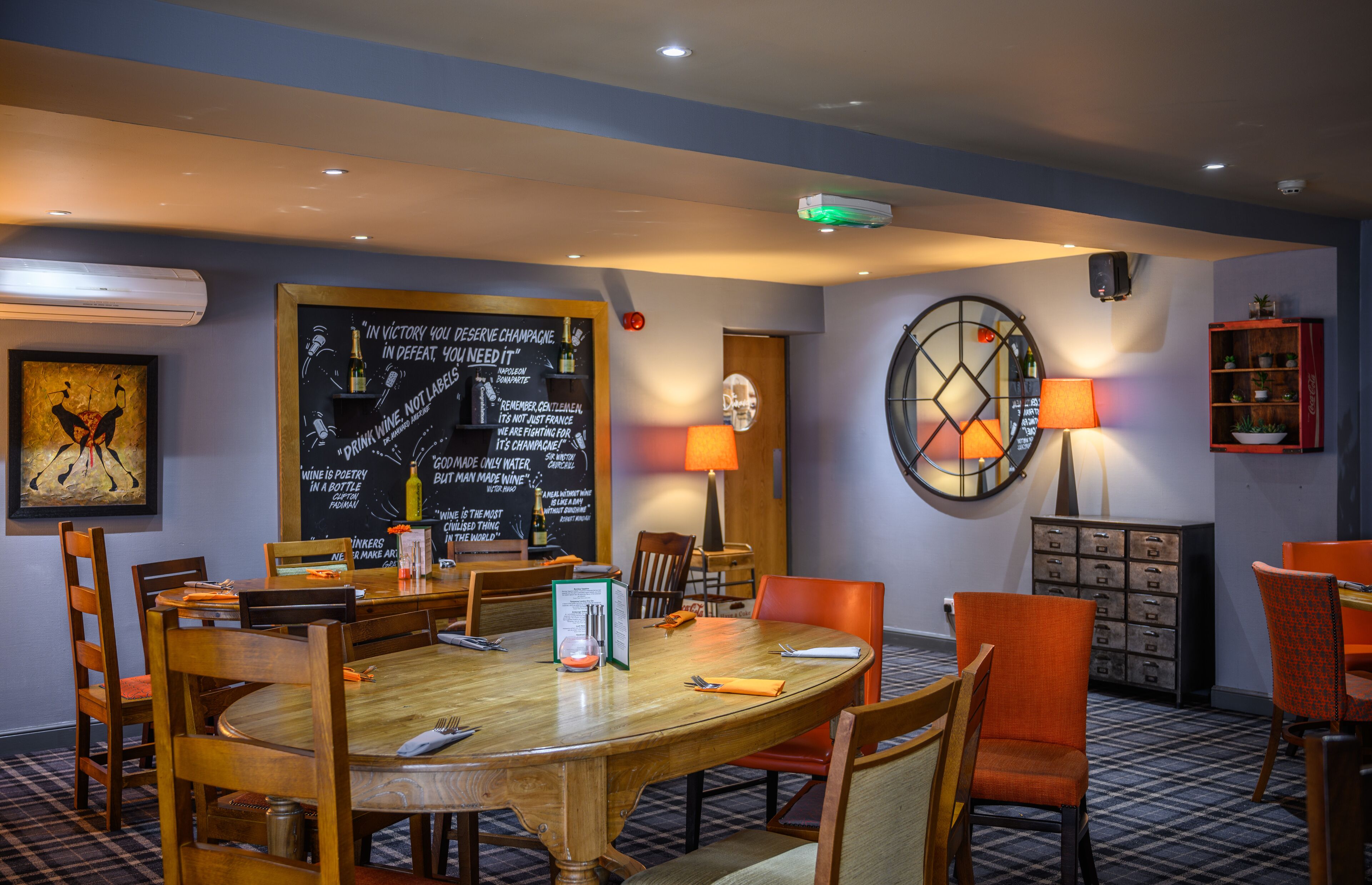 A dining area with a wooden table, chairs, a chalkboard with text, a lamp, and a bookshelf.