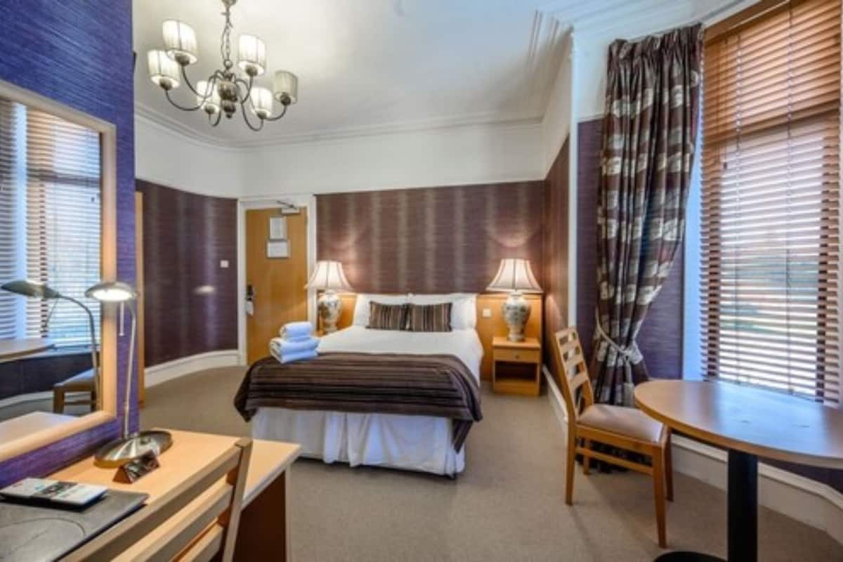A hotel room with a bed, bedside tables, a desk, a chair, a lamp, and a window with curtains.
