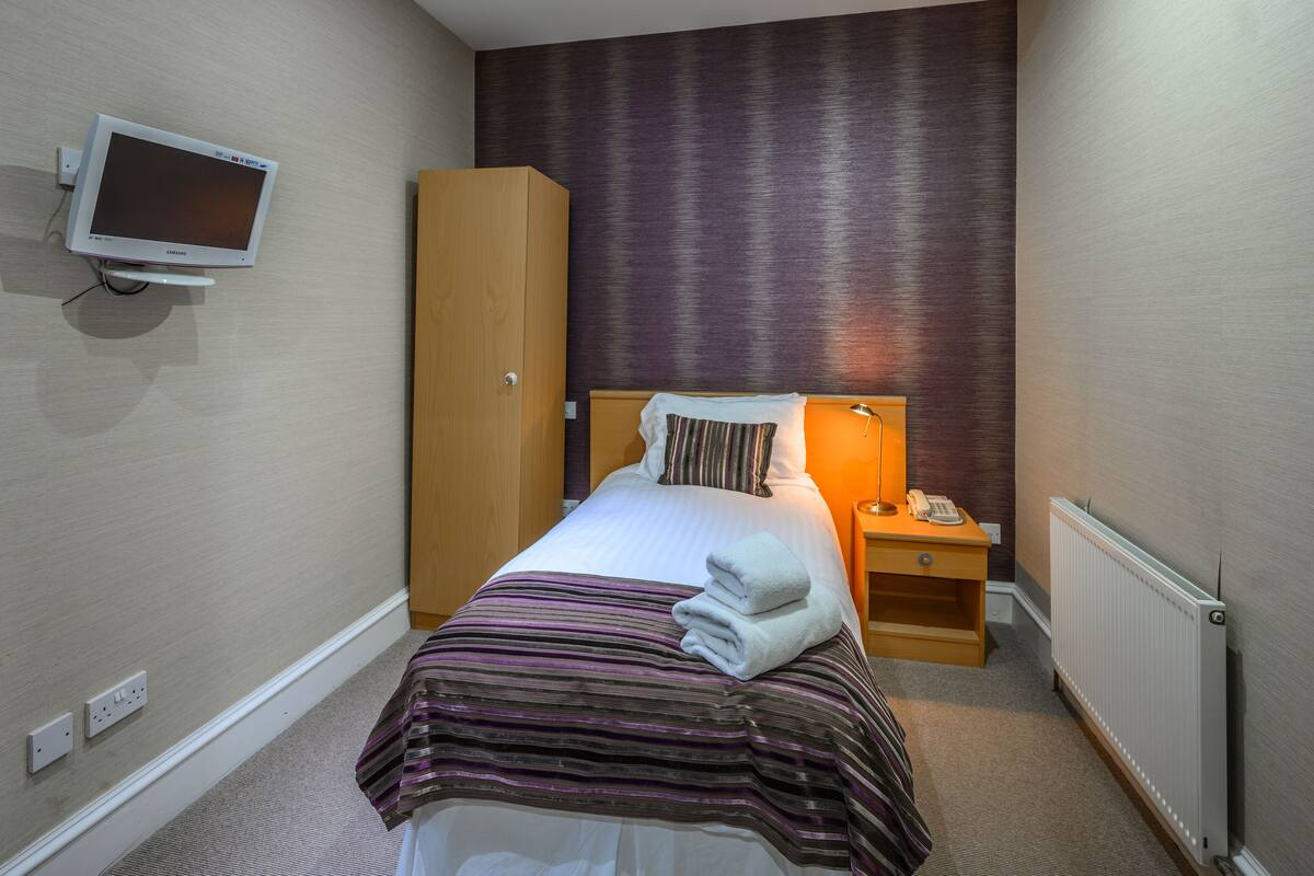A hotel room with a bed, a bedside table with a lamp, a wardrobe, a TV mounted on the wall, and a radiator.