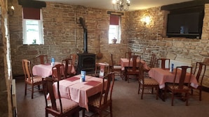 A rustic dining area with stone walls, wooden tables and chairs, a fireplace, and a television mounted on the wall. - Ecclefechan Hotel (Lockerbie)