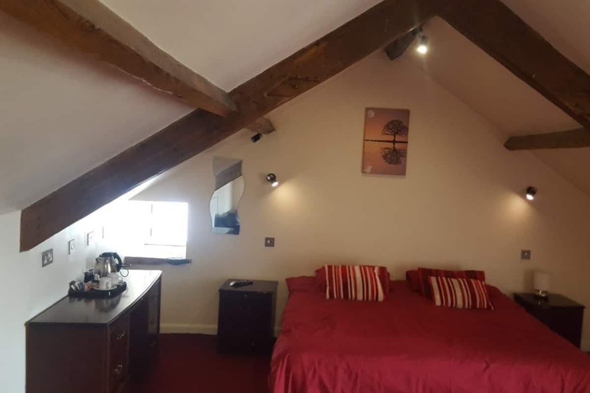 A bedroom with a red bedspread, wooden beams, a painting on the wall, and a desk with a kettle.