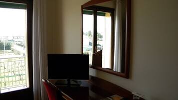 A hotel room with a desk, a television, a mirror, and a window with a view of buildings and greenery.