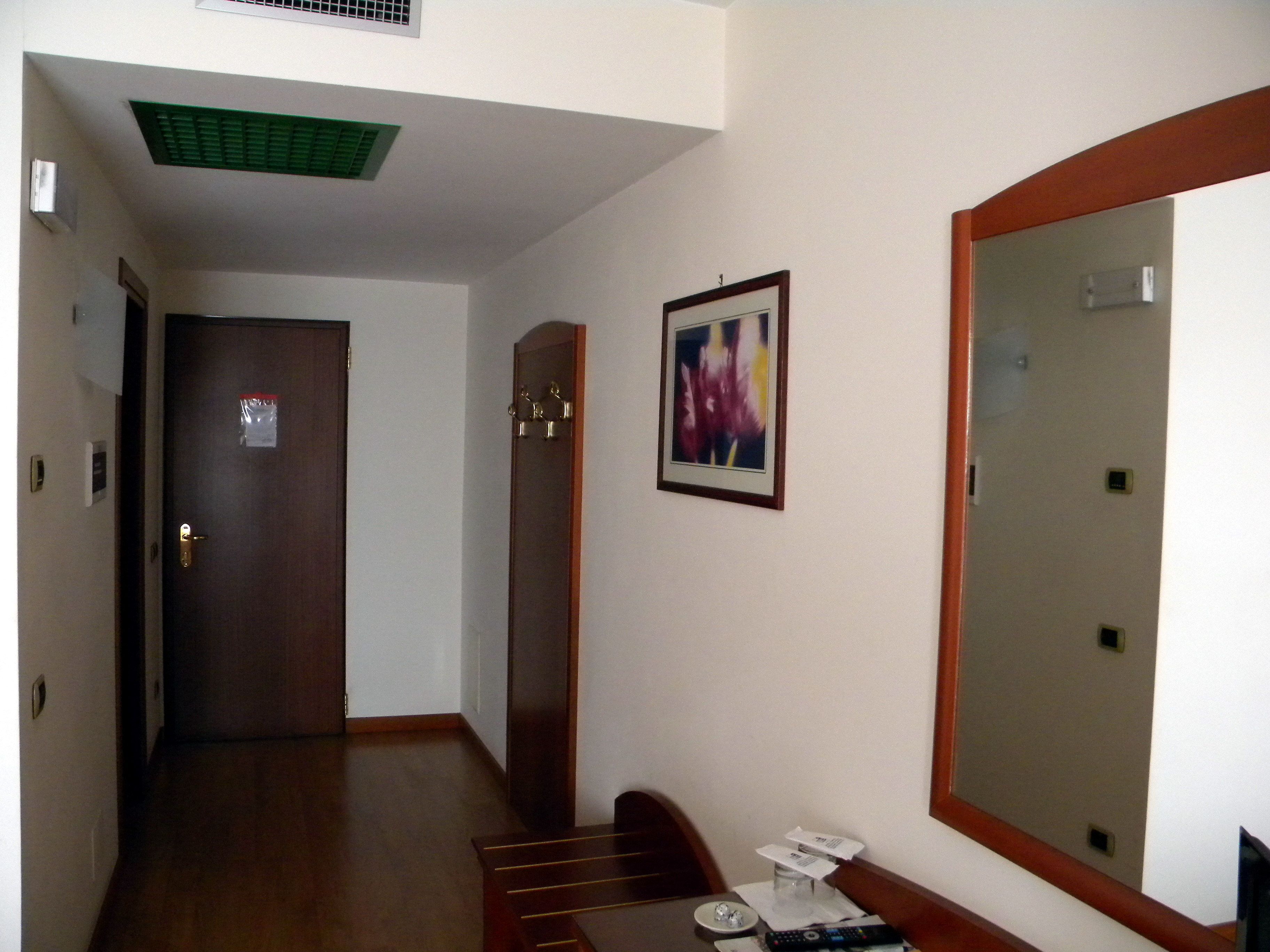 A hallway with a wooden floor, a door with a keyhole, a coat rack, a framed picture on the wall, and a mirror on the right.