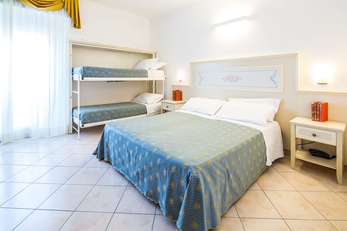 Une chambre d’hôtel avec un lit superposé, un lit simple, une table de chevet avec une lampe, une commode et un tableau encadré au mur.