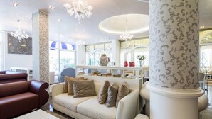 Un salon moderne avec un canapé beige, un divan en cuir rouge et une table basse blanche. On y trouve une grande colonne blanche ornée de motifs, un lustre et un grand vase posé sur une étagère.