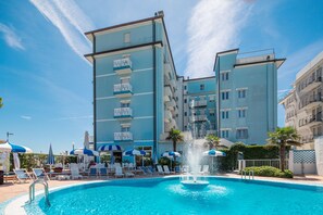 A swimming pool with a fountain, surrounded by lounge chairs and umbrellas, and a multi-story building with balconies. - Hotel Principe (Caorle)