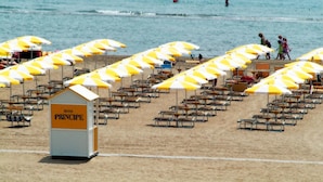 Une plage bordée de rangées de parasols et de chaises de plage aux rayures jaunes et blanches, une tour de surveillance avec l’inscription « Hôtel Principe », et une mer calme en arrière-plan.