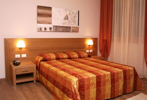 A hotel room with a wooden headboard, two bedside lamps, a telephone, a small bedside table, a bed with a patterned bedspread, a painting on the wall, and a window with curtains. - Hotel Calinferno (Codogne)