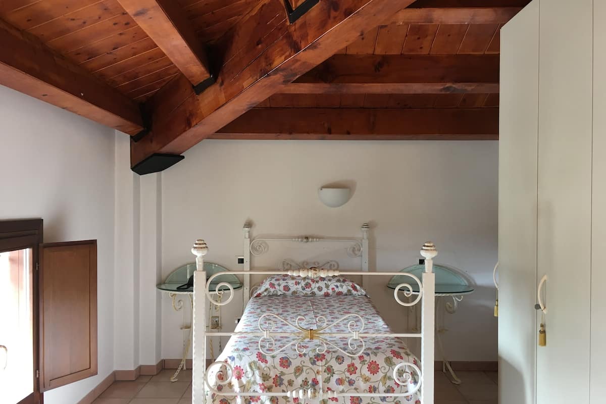 Een slaapkamer met een houten plafond, een bed met een bloemenprint beddegoed, een nachtkastje en een wandlamp.