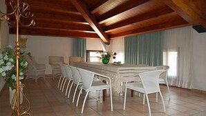 Comedor con una larga mesa y sillas blancas, un candelabro y vistas al exterior a través de amplios ventanales.