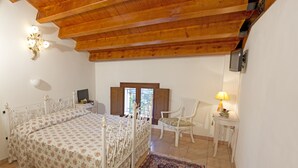 Un dormitorio con techo de madera, una cama con colcha floral, una silla, una mesita con lámpara y una alfombra.