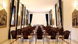 Un salón de conferencias con filas de sillas orientadas hacia una larga mesa, flanqueado por cortinas y obras de arte enmarcadas.