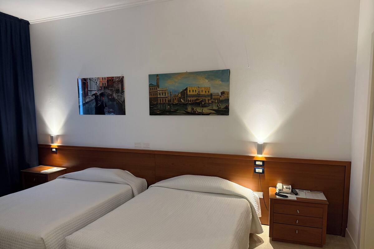 A hotel room with two single beds, a wooden headboard, a nightstand with a lamp, and two paintings on the wall.