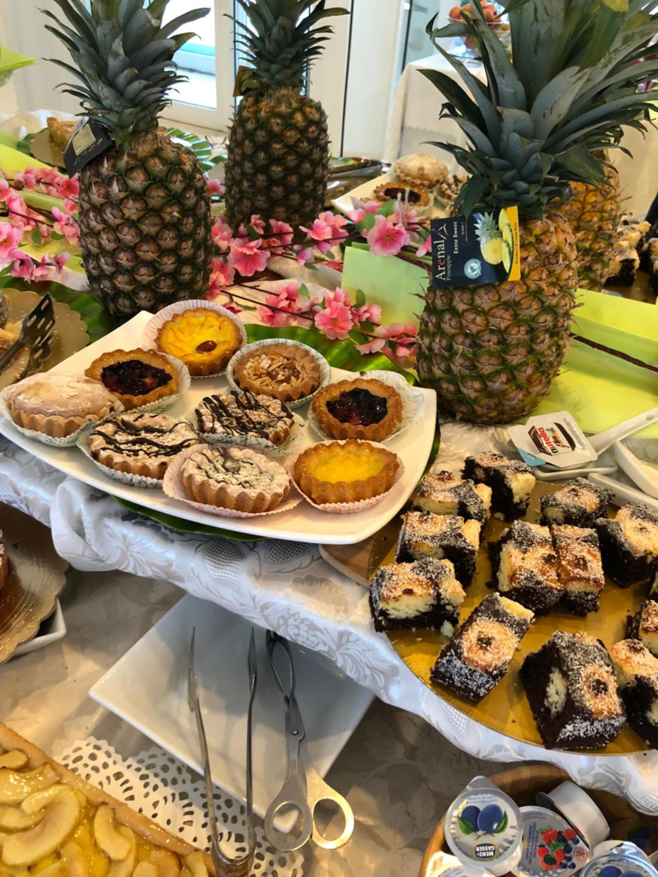 Una mesa de bufé con diversos postres, como tartas, pasteles y productos de pastelería, rodeada de piñas y flores.