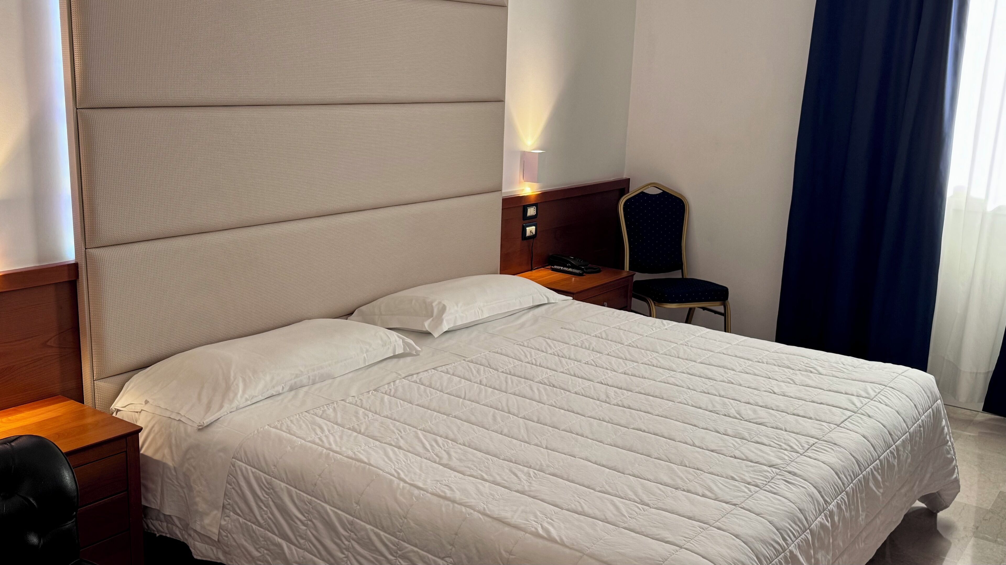 En pænt redt seng med et hvidt dynebetræk, et trænatbord med en lampe og en stol med en mørk pude.