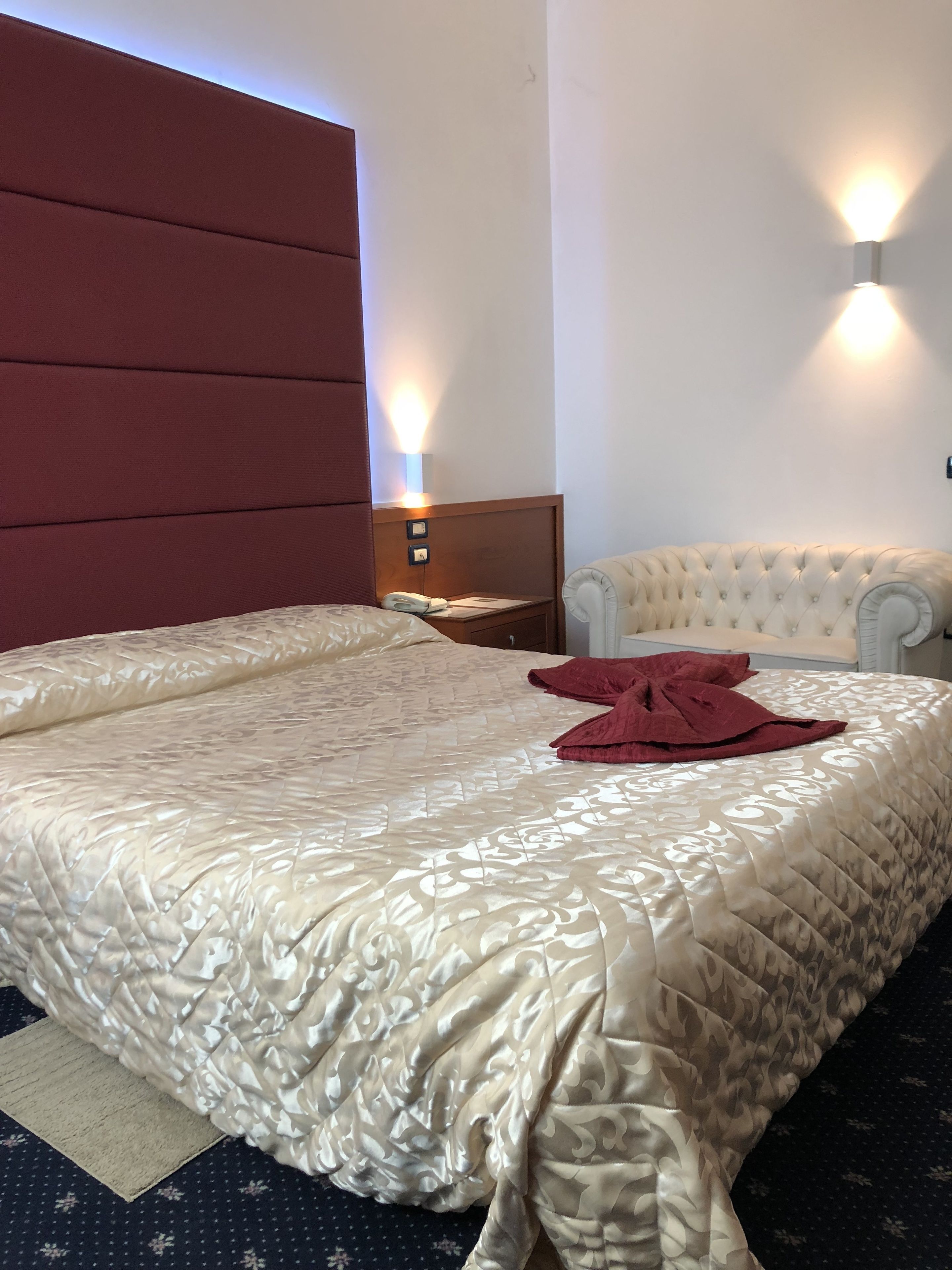 Une chambre d’hôtel avec un grand lit, une tête de lit rouge, une table de chevet avec une lampe et un petit fauteuil.