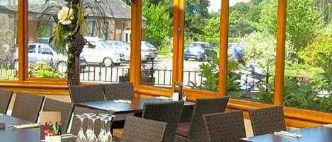 A restaurant with tables and chairs arranged for dining, large windows with a view of parked cars and greenery outside.