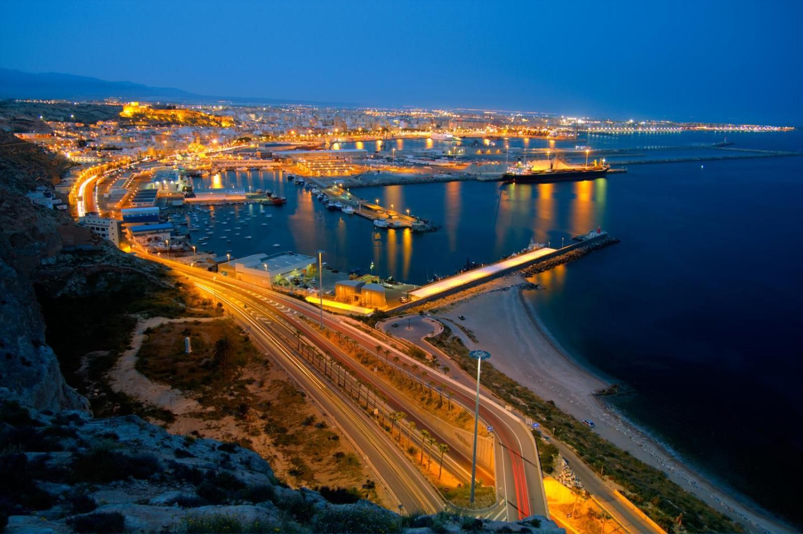 Un paisaje urbano costero al atardecer con calles iluminadas, un puerto deportivo y un puente.