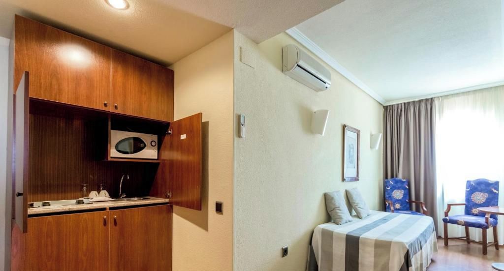 Habitación de hotel pequeña con kitchenette, una cama con colcha a rayas, una silla y una ventana con cortinas.