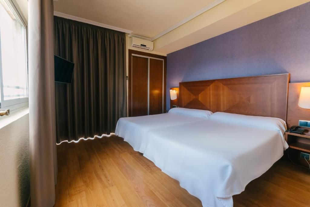 Una habitación de hotel con piso de madera, cama con ropa blanca, cabecera con lámpara y ventana con cortinas.
