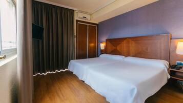 Una habitación de hotel con piso de madera, cama con ropa blanca, cabecera con lámpara y ventana con cortinas.