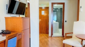 A hotel room with a wooden desk, a flat-screen TV, a telephone, a chair, and a glass shower enclosure. - Apartamentos Torreluz (Almeria)