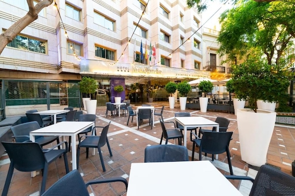 Un área de patio al aire libre con mesas y sillas, un edificio con un letrero que dice 'Hotel Nuevo Horizonte' y banderas.